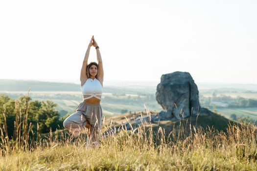 Young Woman Meditating and Practicing Yoga Outdoors at Sunset with Scenic Landscape and Nature Miracle Giant Stone on the Background