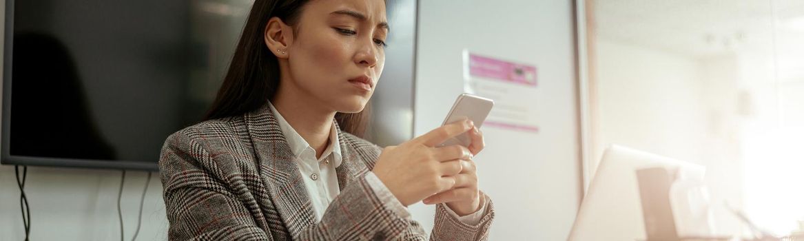 Upset asian business woman using phone while working on her workplace in office