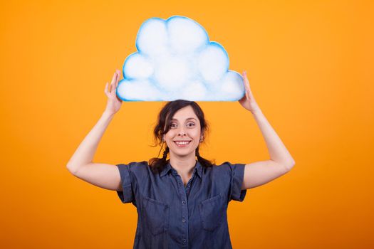 Cheerful young woman smiling at the camera and holding a though cloud on her head in studio over yellow background