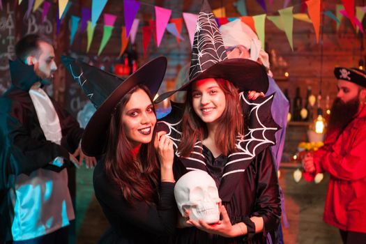Beautiful young witch holding a human skull and smiling to the camera at halloween party
