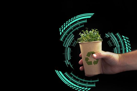 A man's hand holds an eco glass with a green plant on a black background. Blank space for text . Smart and modern recycling. Life without plastic. The concept of ecological technology. Sustainable Development Goals.