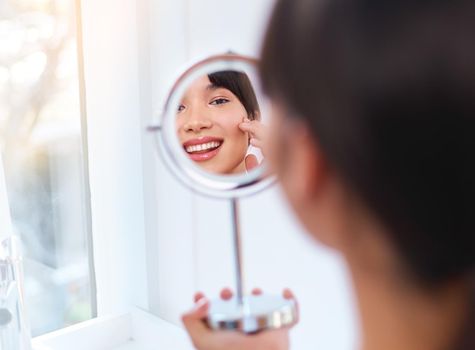 Trying this new stuff out. a cheerful attractive young woman applying moisturizer on her face while looking into her reflexion in the mirror.