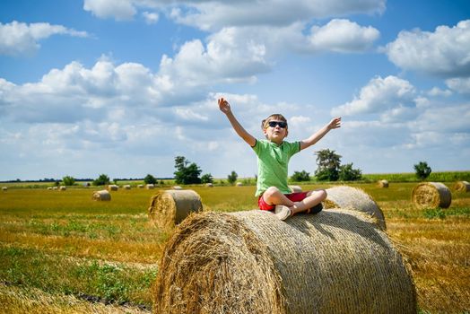 Little boy sits on a round haystack relaxed with hands up. Field