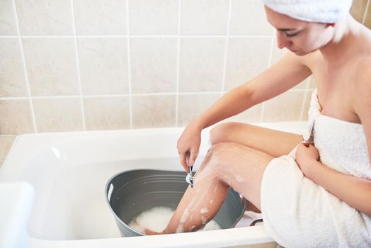 You can definitely still groom yourself and save water. a young woman shaving her legs in a bucket of water in the bathroom at home.