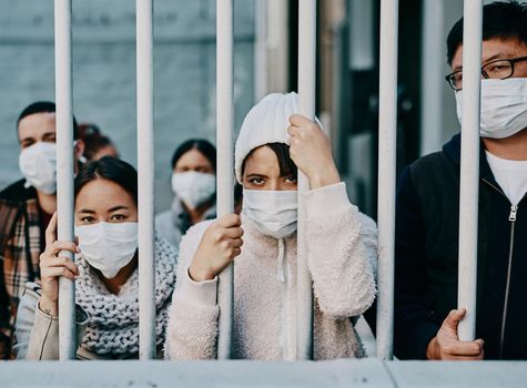 Foreign people in isolation wearing covid face mask at the border or in quarantine or airport looking unhappy, upset and angry. Poor refugees, immigrants and tourists stuck behind a gate in lockdown