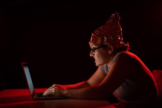 Paranoid caucasian woman typing on a computer wearing a tinfoil cap. Conspiracy theory.
