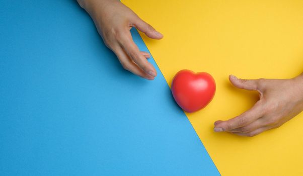 Red heart and stretching hands towards it on a yellow blue background.