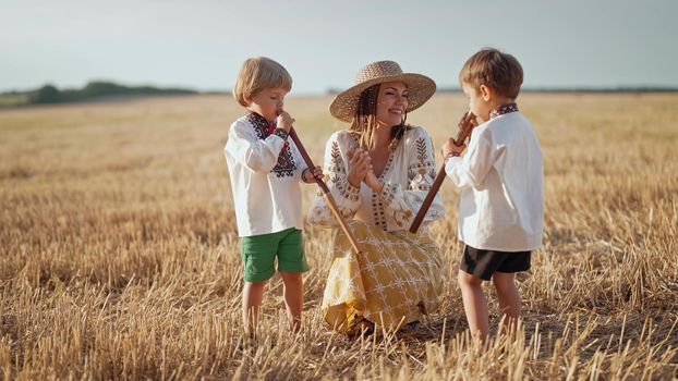 Boys playing on wooden flutes - sopilka. Ukrainian mother with children sons in wheat field. Woman in embroidery vyshyvanka. Ukraine, traditional music instrument, melody, song