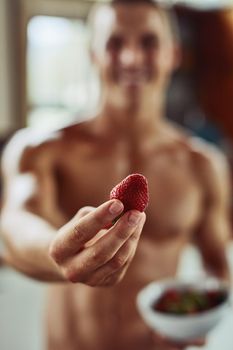 This is for you. Closeup of a cheerful young shirtless man holding out a strawberry at the camera at home during the day.