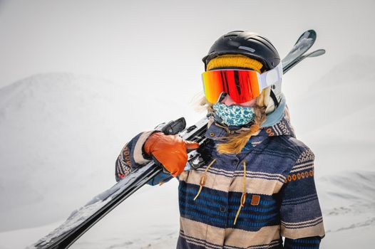 Portrait of a pretty and active woman skier, wearing a mask and holding skis in her hands, active winter holidays