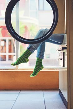 Just minding my own business. an unrecognizable woman seated on a washing machine with her legs showing and showing her bright green striped socks inside of a laundry room.