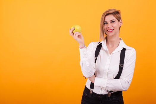 Attractive woman with blond hair keeping arms crossed and holding a green apple over yellow background.