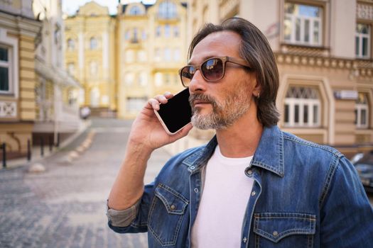 Middle aged man with grey bearded talking on the phone standing outdoors in old city background wearing denim jeans shirt. Mature business man working on the go. Freelancer traveling man