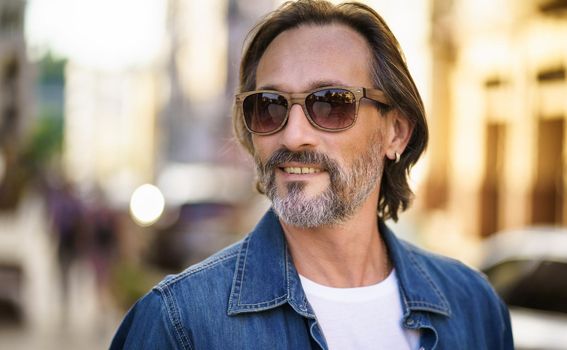 Happy senior man on street of European city wearing sunglasses and denim shirt. Grey bearded Middle aged handsome freelancer man smiling looking sideways wearing casual. Travel concept
