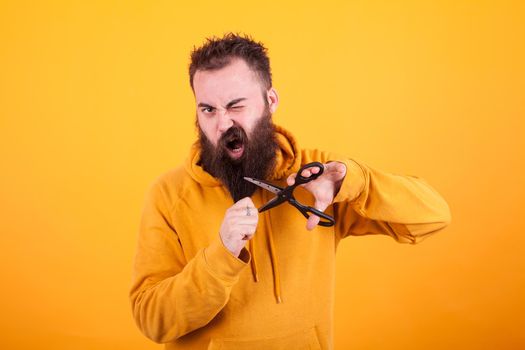 Cool bearded man looking terrified while cutting his beard over yellow background