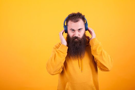 Handsome bearded man looking at the camera and listening music on blue headphones over yellow background
