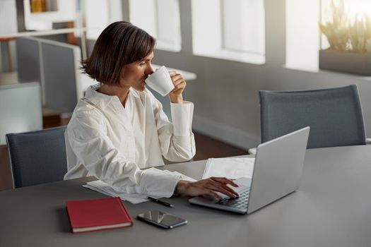Charming businesswoman working at the office using laptop and drink coffee