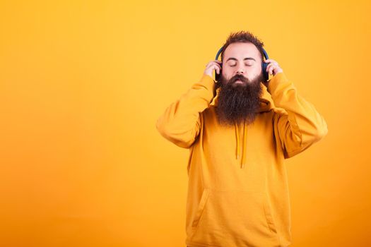 Bearded young man wearing yellow hoodie ,listening music on his blue headphones peacefully over yellow background