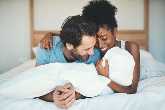 Fun interracial couple laughing, bonding and lying on bed in a home bedroom and looking happy, in love and playful. Smiling man and woman hugging and cuddling after waking up in the morning together