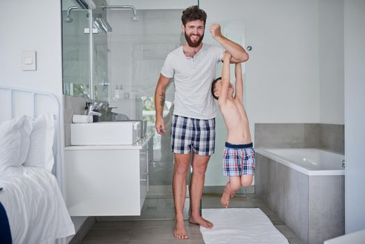 We have tons of fun, from morning till night. a little boy hanging on his fathers arm in the bathroom at home.