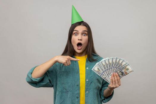 Excited rich woman in party cone on head pointing at money, looking suprised by lottery win.