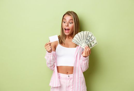 Portrait of attractive rich woman with blond hair, holding money and credit card and smiling amazed, standing over green background