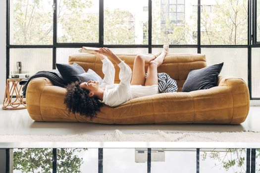 Young happy multiracial woman lying on sofa with legs up, reading a book.
