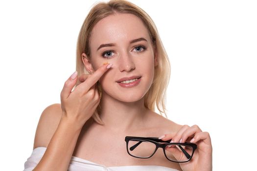 Young blonde woman holding contact lens on finger in front of her face and holding in her other hand a black glasses on white background.