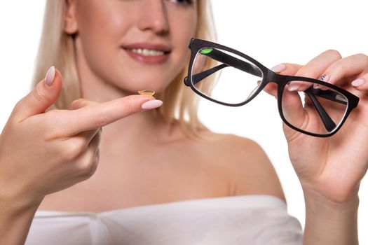 Young blonde woman holding contact lens on finger in front of her face and holding in her other hand a black glasses on white background. Close-up