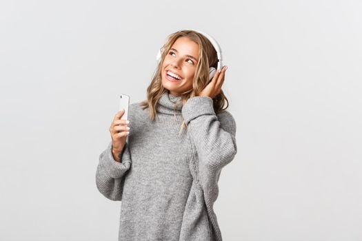 Young attractive blond girl listening music in wireless headphones, smiling and looking at upper left corner, holding mobile phone
