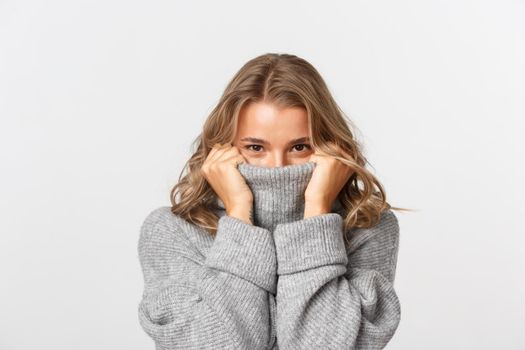 Close-up of beautiful blond girl, pulling sweater on face and smiling, standing over white background