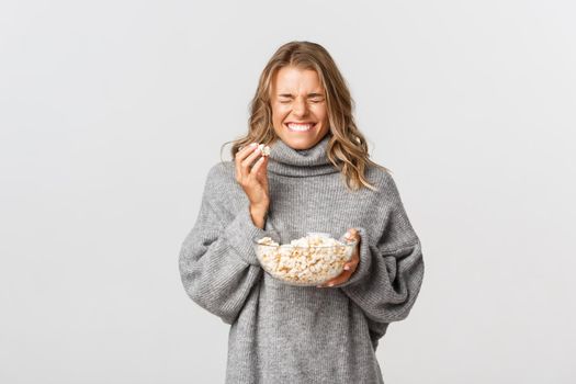 Portrait of happy blond girl, eating popcorn and laughing, watching movie, standing over white background