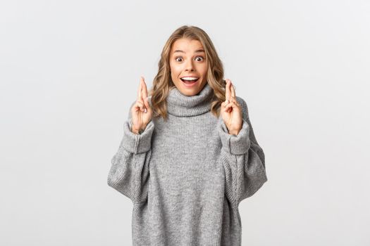 Image of excited and hopeful girl making wish, crossing fingers for good luck, smiling while anticipating results, standing over white background