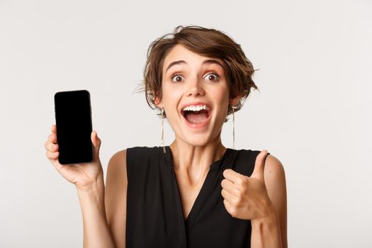 Cheerful beautiful girl showing thumbs-up and demonstrating smartphone screen, standing over white background
