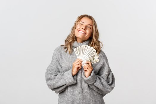 Cheerful blond girl in grey sweater, smiling pleased, holding money, standing over white background