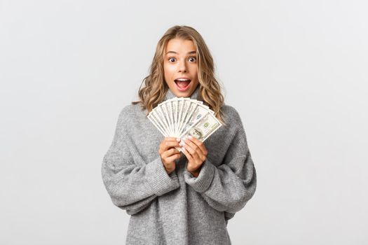 Image of excited attractive girl in grey sweater, holding money and looking amazed, standing over white background