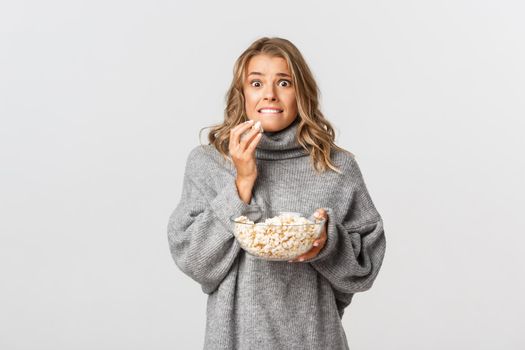 Portrait of blond girl eating popcorn and looking scared, watching movie, standing over white background