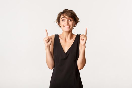 Hopeful girl pointing fingers up and smiling, looking at camera excited, standing over white background