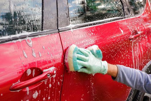 Worker washing red car with sponge on a car wash.male hand holds with green red sponge washing car. Concept car wash clean.Washing with soap.