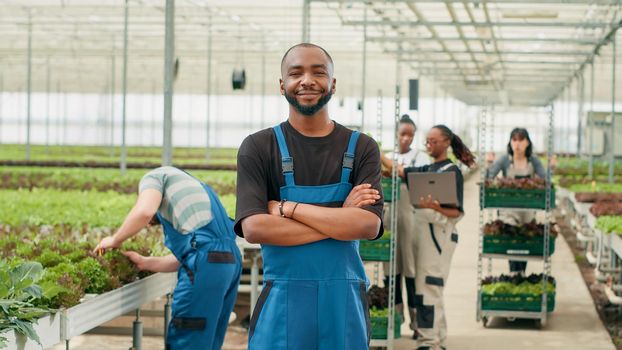 Smiling african american man posing with arms crossed while farm workers using laptop manage deliveries