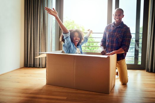 Surprise everyone. a cheerful woman inside of a cardboard box with her boyfriend standing next to her inside at home during the day.