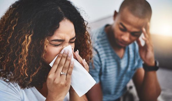 Flu season is here. a young woman blowing her nose with her boyfriend looking irritated in the background.