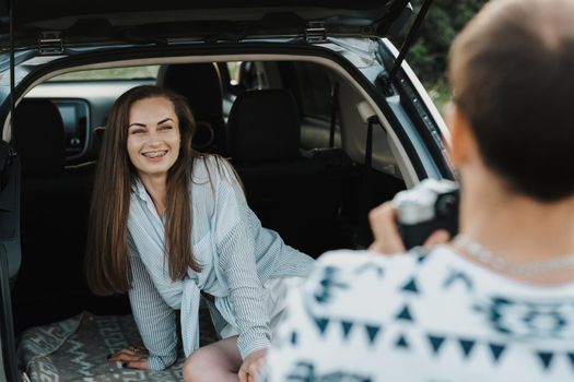 Cheerful caucasian woman sitting in trunk of SUV car while boyfriend photography her on a camera