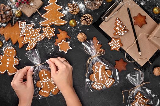 Christmas gift gingerbread on dark background. Biscuits in festive packaging. Woman is packaging Christmas gingerbread cookies with icing sugar. Top view