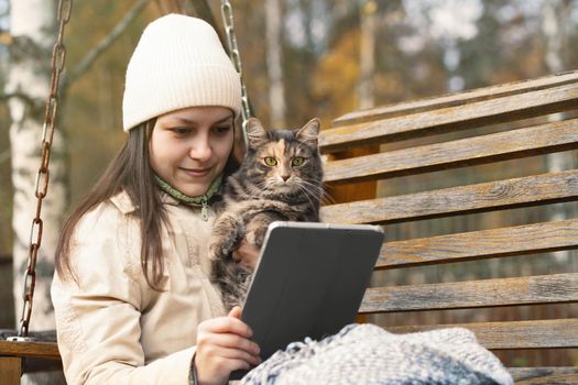 Girl uses a tablet while sitting on a bench in an autumn park with a cat in her arms