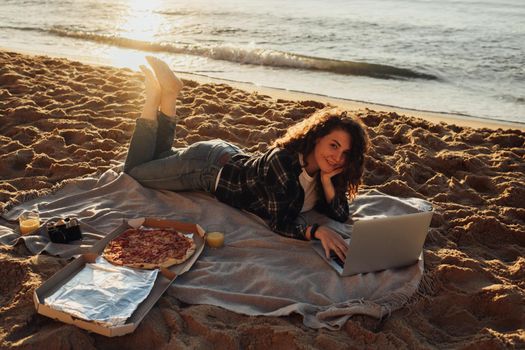 Happy curly haired woman eating pizza and working on laptop while laying on the seashore at dawn