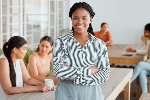 Motivated, ambitious and confident young business woman standing arms crossed in the office break room with colleagues in the background. Portrait of a female leader feeling happy and positive