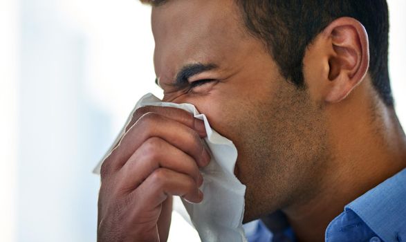 Struggling to fend off the flu this season. a young businessman blowing his nose at work.