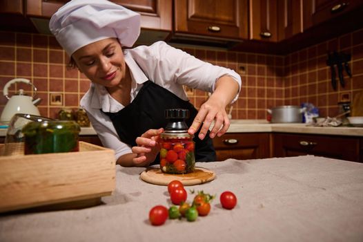 Beautiful Hispanic housewife using special key, closes lid of jar filled with ripe and juicy tomatoes while canning