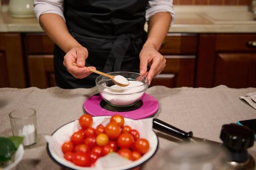 Details: housewife's hand holds wooden teaspoon, weighing sugar on kitchen scale while marinating and canning tomatoes